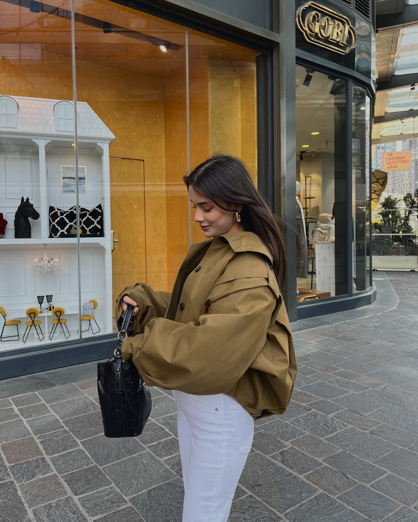 vrouw draagt camel cropped trench jacket met zwarte top en witte broek, zijaanzicht