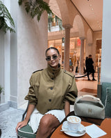 vrouw in camel cropped trench jacket met opstaande kraag en plissérok in chic café
