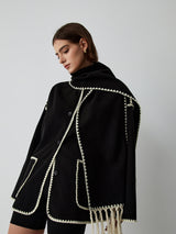zwarte cape-jas met geïntegreerde sjaal en contrasterende bies, gedragen door model