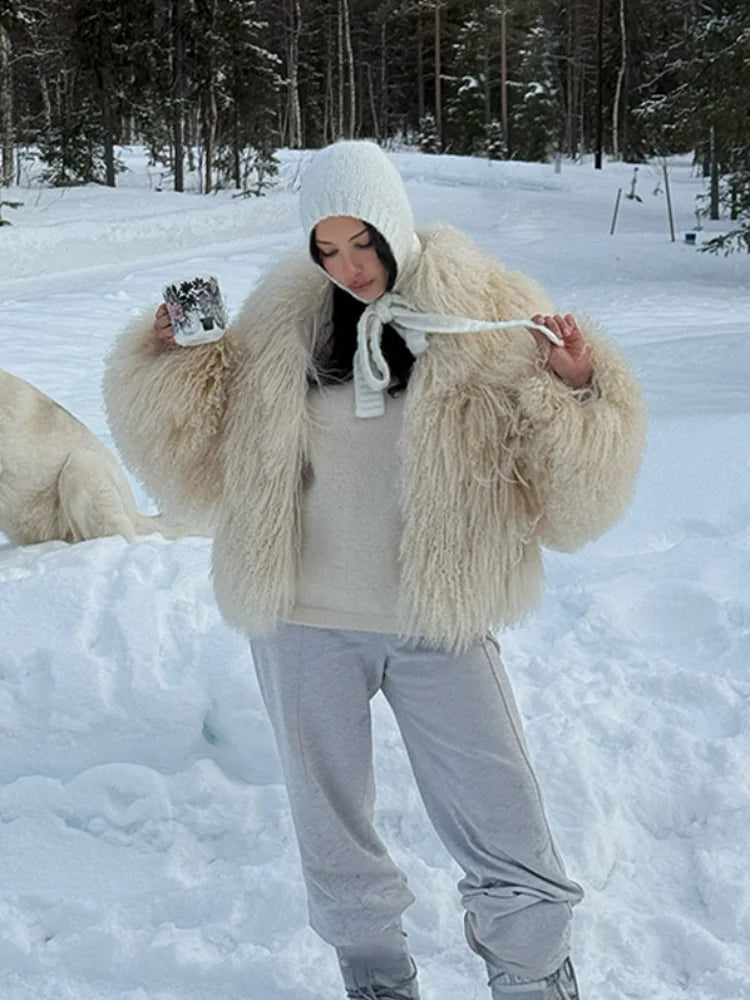 vrouw draagt beige fluffy jas in de sneeuw met witte broek en laarzen