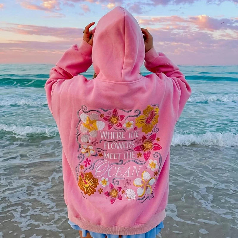 roze hoodie met geborduurde bloemen en tekst 'where the flowers meet the ocean' op het strand